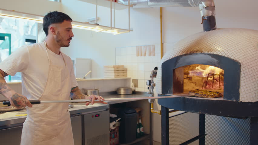 Male chef in apron putting hot pizza on metal plate after baking, using peel to take it off wood-fired oven at work in pizzeria kitchen