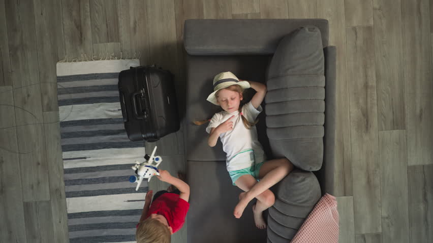 Tired girl waves hand imitating conducting orchestra on sofa while toddler playing with airplane. Children take break after long flight upper view Royalty-Free Stock Footage #3424429465