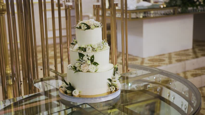 A three-tiered wedding cake adorned with white roses and green foliage, displayed on a mirrored table with golden support bars. The cake's elegance is accentuated by the luxurious surroundings