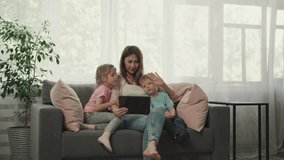 Mother and children talk to father on video call via tablet waving hello with hands. Happy family enjoys online communication sitting on sofa