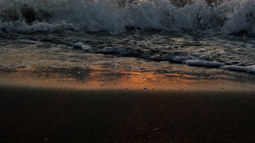 Sea waves gently pour over the black sand shore and the golden setting sun is reflected on the water. A sea wave washing the shore of a beach with volcanic sand and the red reflection of the sunset