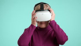 Impressed elderly woman with short blonde hair, light make up in purple turtleneck trying on VR headset, pointing fingers, hands in space, blue background. High quality 4k footage - Powered by Shutterstock - Get 15% off with code: PIKWIZARD15