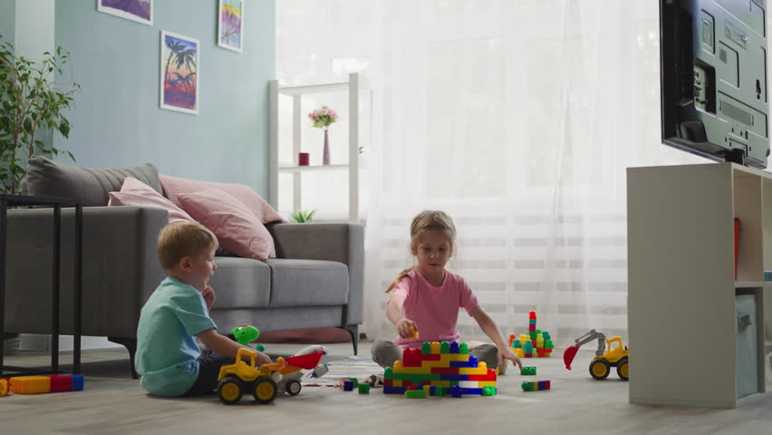 Toddler boy plays with airplane while preschooler builds wall from colorful constructor blocks. Girl enjoys creative activity sitting on floor with brother Royalty-Free Stock Footage #3424560477