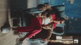 Young girl wearing fur coat with leo print and sunglasses sitting cross-legged in old armchair and posing on camera with confidence in room with antique interior. Video portrait, vertical clip