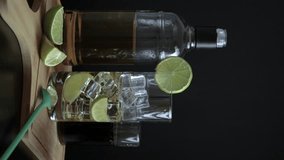 Piscola, preparing, cocktail, hand adding cola, glass with ice, wooden board, slice of lemon, spoon. On wooden board and black background. Original from Chile, summer, video, vertical. Typical Chilean