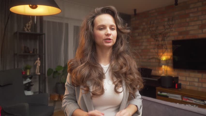 A woman with a beautiful voluminous hairstyle on long curly brown hair communicates and explains something, actively gesticulating with her hands during a conversation from home via online video link. Royalty-Free Stock Footage #3424690379