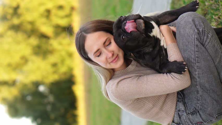 Woman hugs her adopted black French bulldog tightly during a walk in the park. The concept of helping homeless animals, animal adoption, friendship, trust, love, care. Vertical Royalty-Free Stock Footage #3424720049
