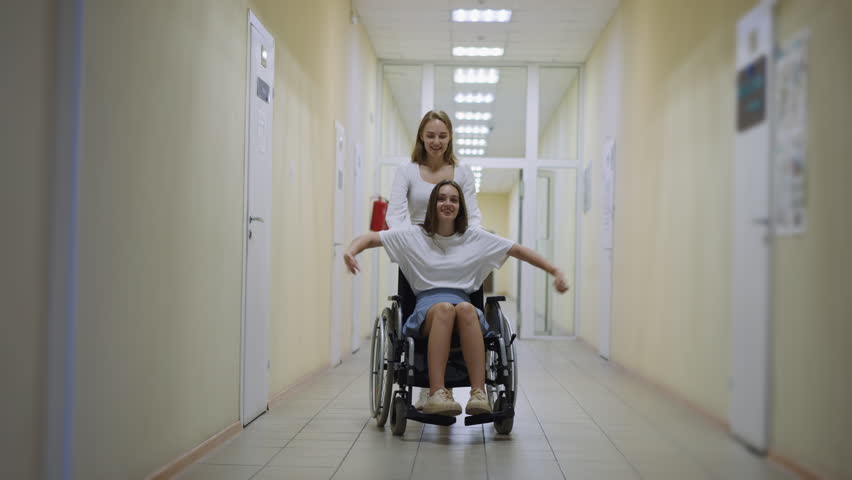 Happy woman runs pushing wheelchair with disabled sister laughing and spreading hands. Cheerful ladies have fun after therapist appointment Royalty-Free Stock Footage #3424727965