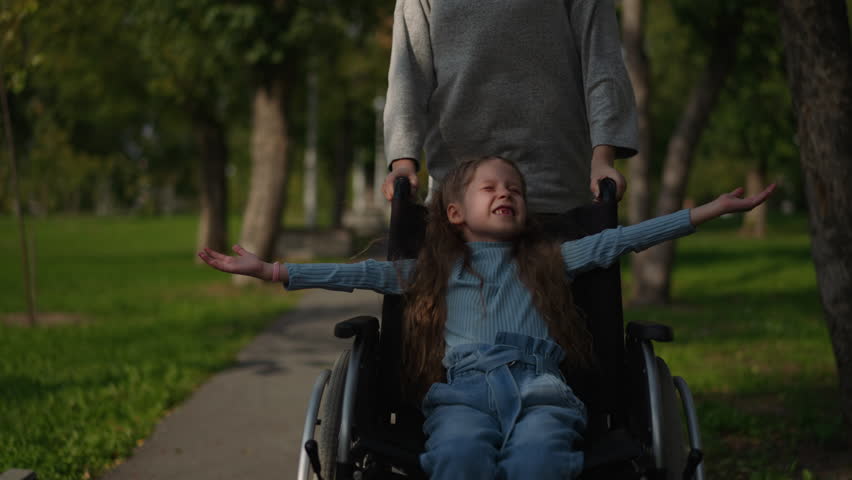 Toothless little girl smiles in wheelchair spreading hands to sides. Mother pushes medical vehicle with child running on road in spring park closeup Royalty-Free Stock Footage #3424736605