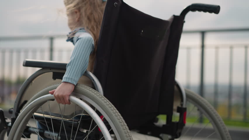 Little girl turns wheels of medical equipment to move on city viewpoint. Schoolchild with chronic health condition controls wheelchair closeup Royalty-Free Stock Footage #3424760547