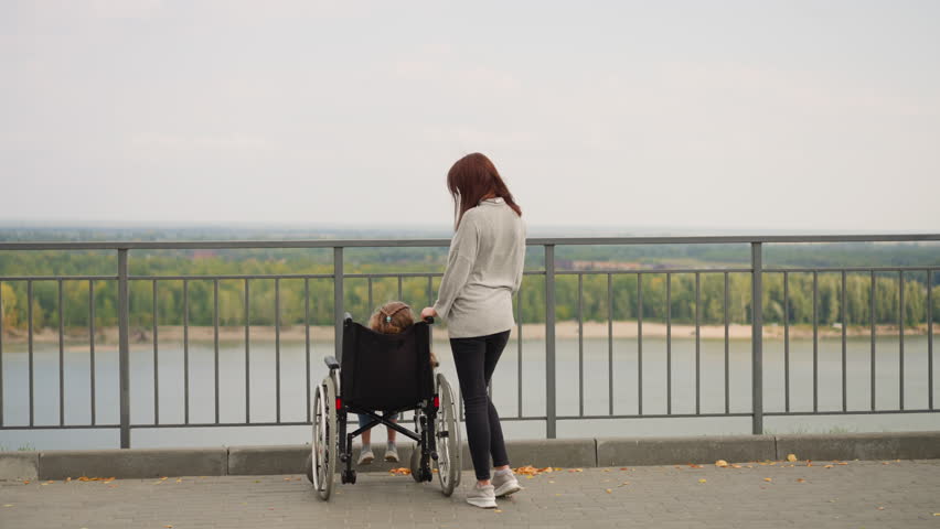Woman shows large park with green trees to daughter with spinal cord injury. Mother and little girl in wheelchair on observation deck overlooking river