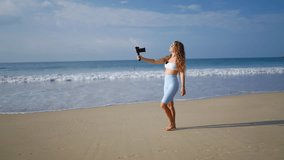 Fashionable female vlogger records content with gimbal stabilized camera at beach. Blonde influence creates travel vlog, walking barefoot on sandy shore. Videography of ocean waves, tropical scenery.