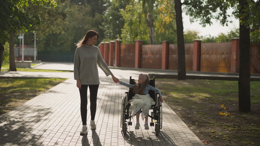 Happy woman and little girl join hands enjoying walk in city park on sunny spring day. Loving mother looks at daughter sitting in wheelchair Royalty-Free Stock Footage #3424775535