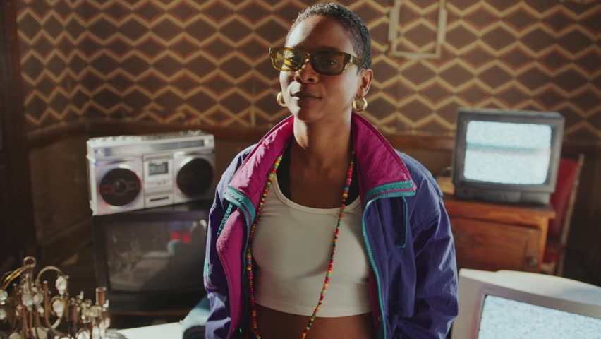 Stylishly dressed Black girl in 80s shell suit moving her head to the beat and enjoying the music in old room, analog TVs with no signal screen and boombox in the background