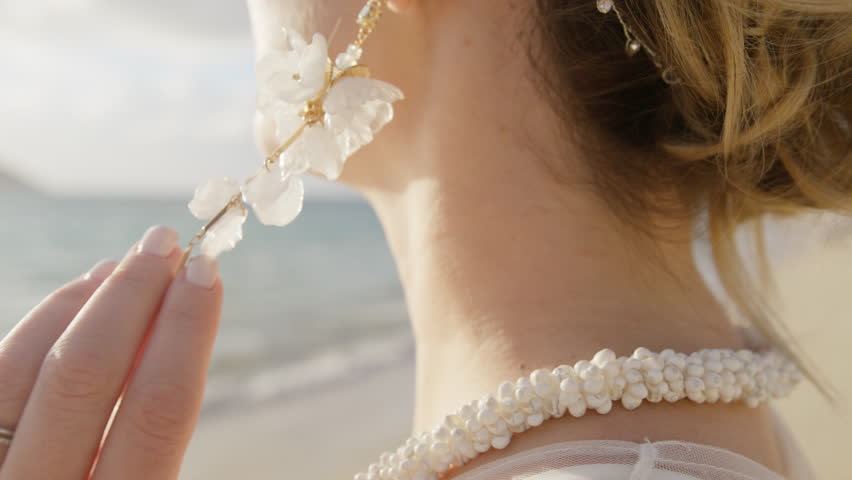 Side view of bride slowly touching boho style floral drop earrings. Girls hand with wedding diamond ring at beach 4K. Close up side view of beautiful woman wearing luxury hanging white flower earrings