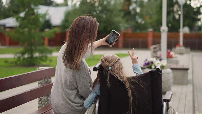 Mother makes fun selfies with little daughter in wheelchair sitting in summer park. Girl shows finger gestures in camera of smartphone backside view
