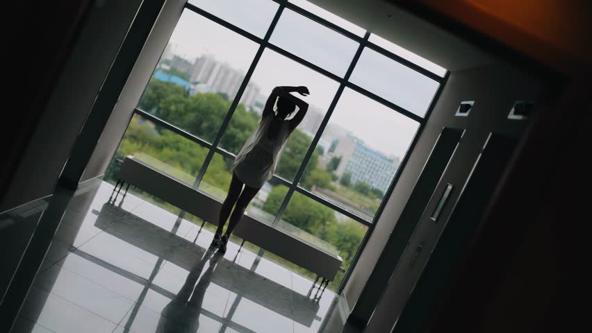 The girl is standing at the end of the corridor in front of a tall bright window. She is wearing a light negligee. Shooting from behind.