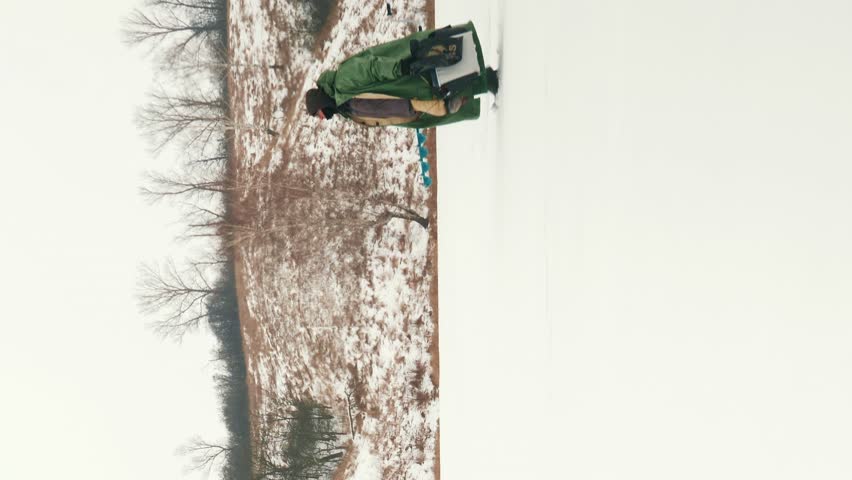 Fisherman with fishing equipment and ice drill is going through frozen lake. Man wearing winter extreme cold clothing is walking along frozen river in winter. Vertical video