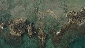 Drone footage: top-down view of a rocky reef surrounded by vibrant blue waters, with dynamic whitewash gracefully dancing around the rocks