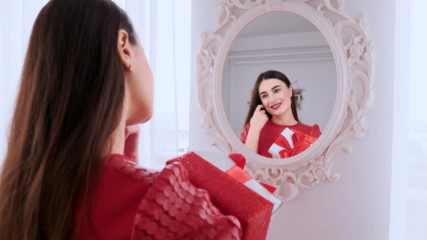 Beautiful happy girl dressed in red dress, smiles and looks in the mirror. She holds the gift boxes. Valentine