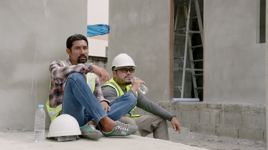 Two male construction engineers wearing hard hats, reflective vests, for safety when working in construction zone, sit rest during the day from construction work, drink water relieve their fatigue.