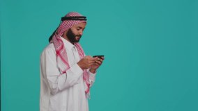 Islamic guy playing fun mobile videogames on phone, dressed in traditional gown with headscarf. Middle eastern person engaging in online gaming contest using smartphone app.