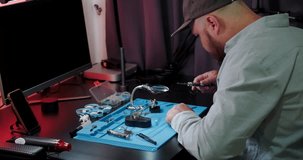 Medium shot of a man sitting at his desk in a shirt, soldering a video transmitter chip from an FPV drone. A young engineer solders an electronic board. In the evening the man is busy with his hobby.