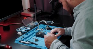 Medium shot of a man sitting at his desk in a shirt, soldering a video transmitter chip from an FPV drone. A young engineer solders an electronic board. In the evening the man is busy with his hobby.