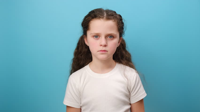 Portrait of worried little girl kid covering mouth with hand, afraid to say secret, child terrified to speak, wearing white t-shirt, posing isolated over plain blue color background wall in studio Royalty-Free Stock Footage #3424977835