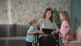 Mother smiles showing videos to curious children on tablet. Toddler and preschooler stand near mommy with disability sitting in wheelchair in room