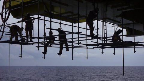 Silhouette of scaffold erector team with falling protection hanging on scaffold frames below oil and gas platform during working overboard in the middle of the sea.