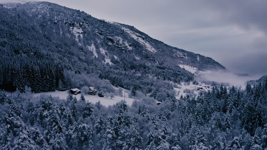 Secluded farm houses in a mountain landscape, winter snow covered forest, low fog covering a village at a distance. Low light slow motion aerial drone footage. Royalty-Free Stock Footage #3425102559