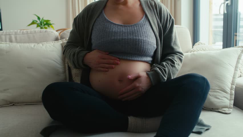 Unrecognizable caucasian pregnant woman sitting on sofa and touching her abdomen