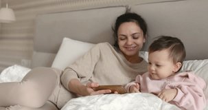 Mother and cute baby playing with smartphone while sitting on bed in bedroom at home