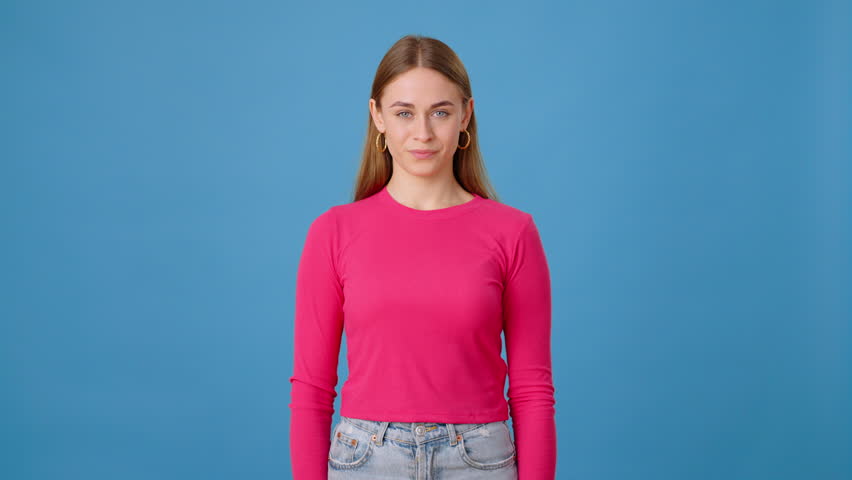Confident lady in pink long sleeve pointing at camera with index finger and clapping with proud face in blue studio. Smiling woman congratulating with applause and expressing approval indoors. Royalty-Free Stock Footage #3425202329