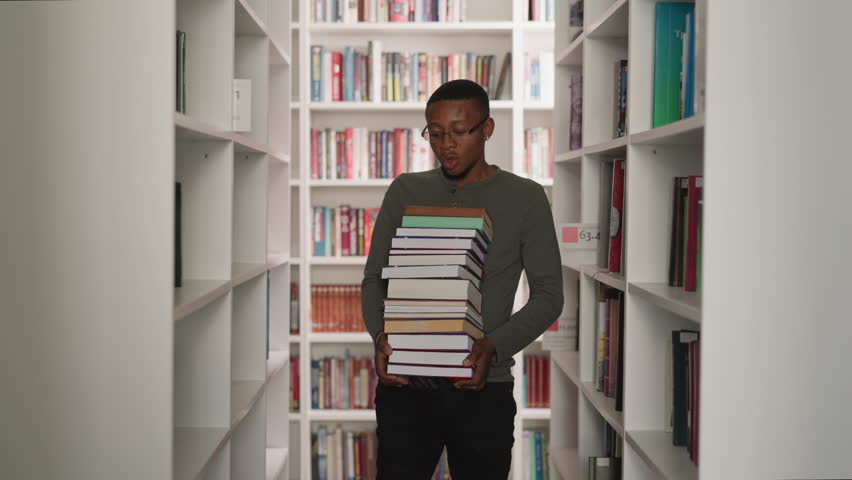 Emotional man carries book stack in library. African American librarian holds large literature pile walking between bookcases. Student in archive Royalty-Free Stock Footage #3425217707