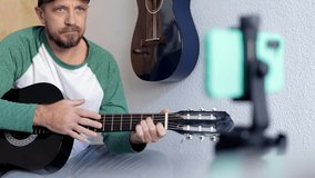 A young European musician teaches guitar lessons online live from a smartphone to a student. 
