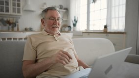 Senior man talking video call on laptop. Old gray haired male in glasses chatting on computer sitting on sofa at home looking at screen. Pensioner enjoying online conversation with family, friends.