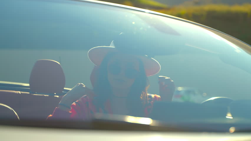 Succesful brunette young woman driver preens her hair sits in convertible car and looks at mirror, view through windshield. Tourism, road trip, journey, vacation, travel, car trip, wealth concept.