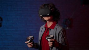 A cheerful teenager in a VR helmet on his head energetically moves controllers in his hands. A teenager stands and smiles while playing a video game in a VR club room