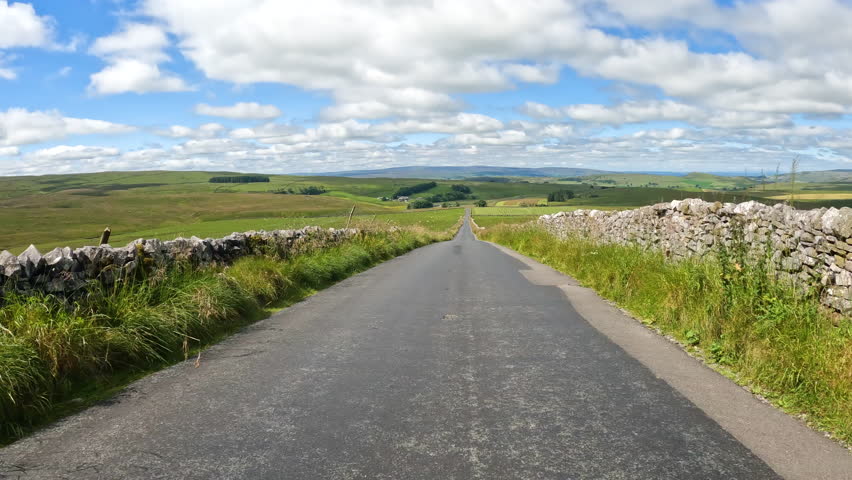 POV: Driving past countless sheep pasturing on wide green meadows of Yorkshire Dales National Park. A beautiful sunny summer day for a road trip through the picturesque green English countryside.
