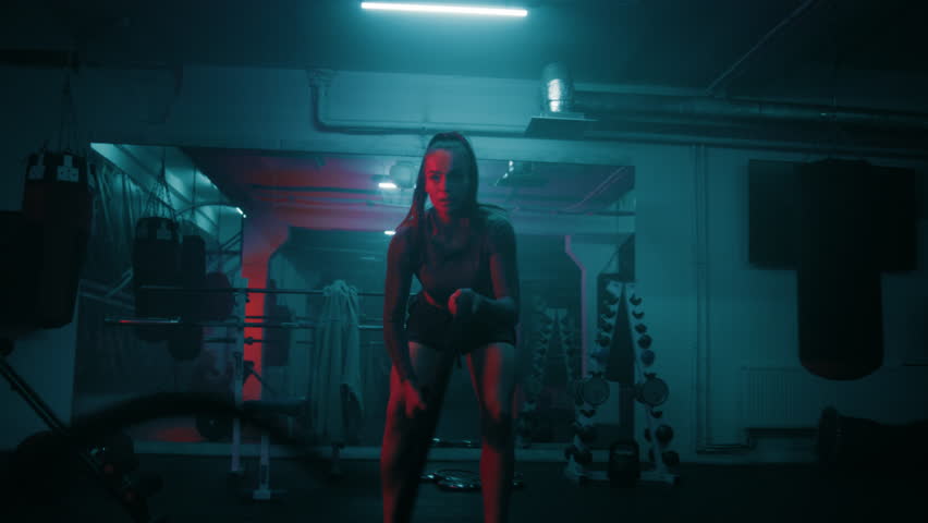 Athletic woman trains with battle ropes in dim boxing gym with LED lighting. Professional female boxer does cardio or endurance exercises before fighting competition. Intensive CrossFit workout.