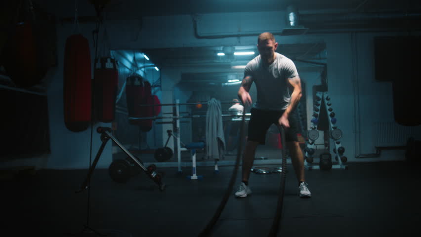 Male athlete exercises with battle ropes in dark boxing gym with LED lighting. Professional boxer does cardio or endurance workout before championship fight. Physical activity and CrossFit training. Royalty-Free Stock Footage #3425466605