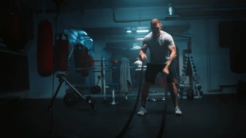 Male athlete exercises with battle ropes in dark boxing gym with LED lighting. Professional boxer does cardio or endurance workout before championship fight. Physical activity and CrossFit training. Arkistovideo