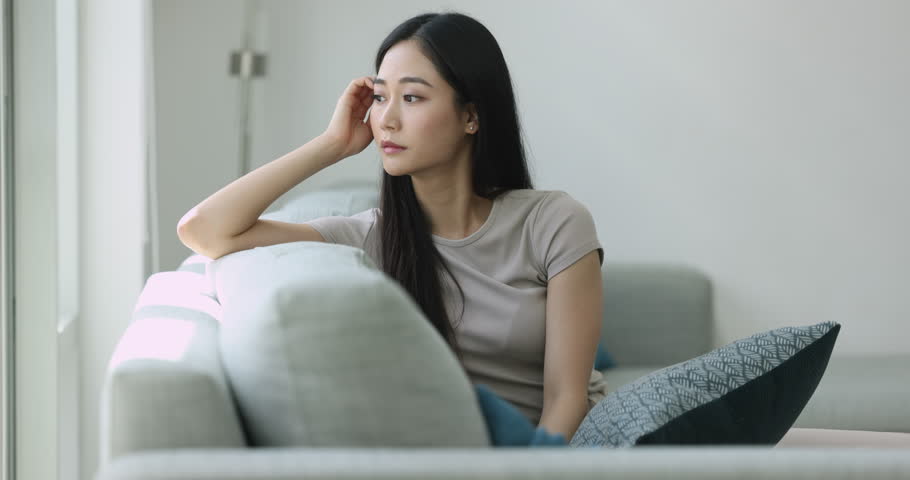 Serious pensive young Asian woman thinking on problems, sitting on soft couch at home, touching head, hair in deep sad thoughts, looking at window away, feeling depressed, frustrated Royalty-Free Stock Footage #3425605075