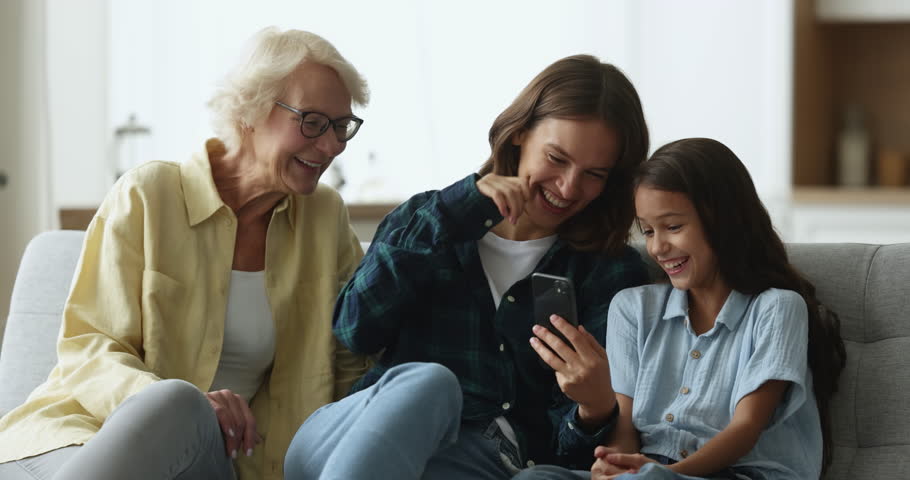 Happy grandma, mother and cheerful girl holding smartphone and laughing out loud, looking at screen, having fun, relaxing on comfortable sofa, using funny media service, online application for selfie Royalty-Free Stock Footage #3425607731