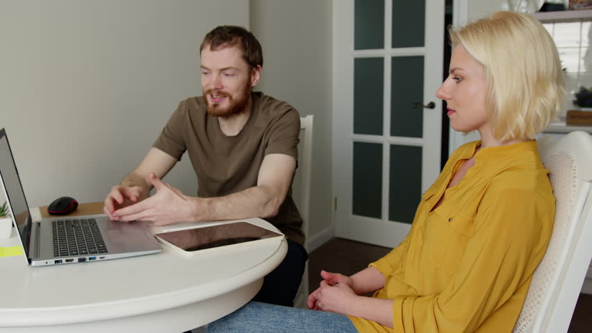 Young blond woman with stylus pen using tablet while sitting at the table with laptop near young bearded man in the kitchen. Royalty-Free Stock Footage #3425611399