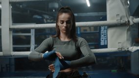 Woman sits near boxing ring, wraps her hands with bandage for martial arts and looks at camera. Athlete prepares to fight, competition or training in box gym. Boxers warm up at background. Portrait. - Powered by Shutterstock - Get 15% off with code: PIKWIZARD15