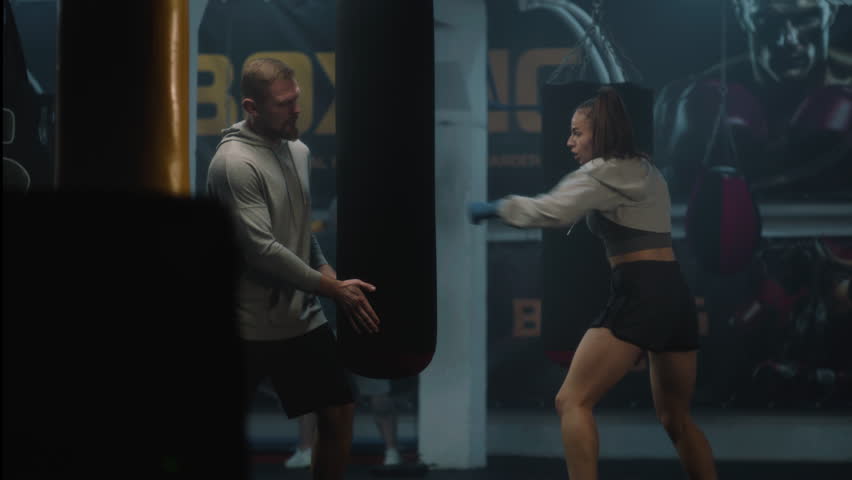 Athletic woman in boxing bandages hits punching bag while training in dark gym. Female kickboxer practices and prepares for competition with male coach. Physical activity and intensive workout.