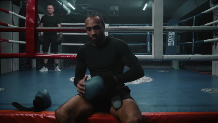 African American fighter sits near ring and puts on boxing gloves. Adult boxer comes in boxing ring and starts to train punches with coach. Athlete prepares to tournament or competition in boxing gym.